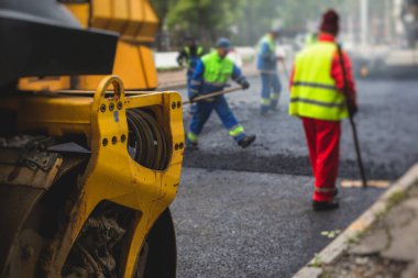 Asfalt işleme, asfalt kaplama ve asfalt kaldırım, arka planda asfalt kaldırım makinesi ve buhar silindirli sıkıştırma aracı ile asfalt tabakasını bitiren bir grup işçi.