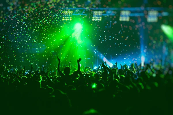 A crowded concert hall with scene stage green lights, rock show performance, with people silhouette, confetti explosion fired on dance floor air during a concert festival