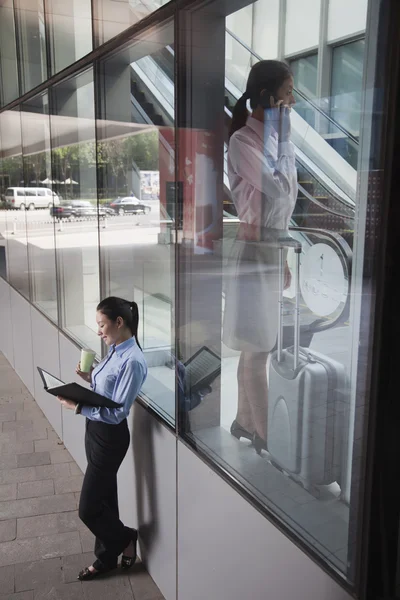 Jonge zakenvrouw in het gebouw praten over de telefoon, een andere vrouwen buiten het gebouw kijken naar haar notities — Stockfoto