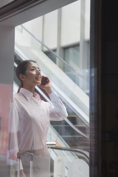 Młoda kobieta wewnątrz budynku, rozmawia przez telefon i patrząc przez szybę — Zdjęcie stockowe