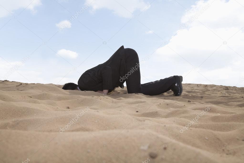 Businessman kneeling with his head in a hole in the sand