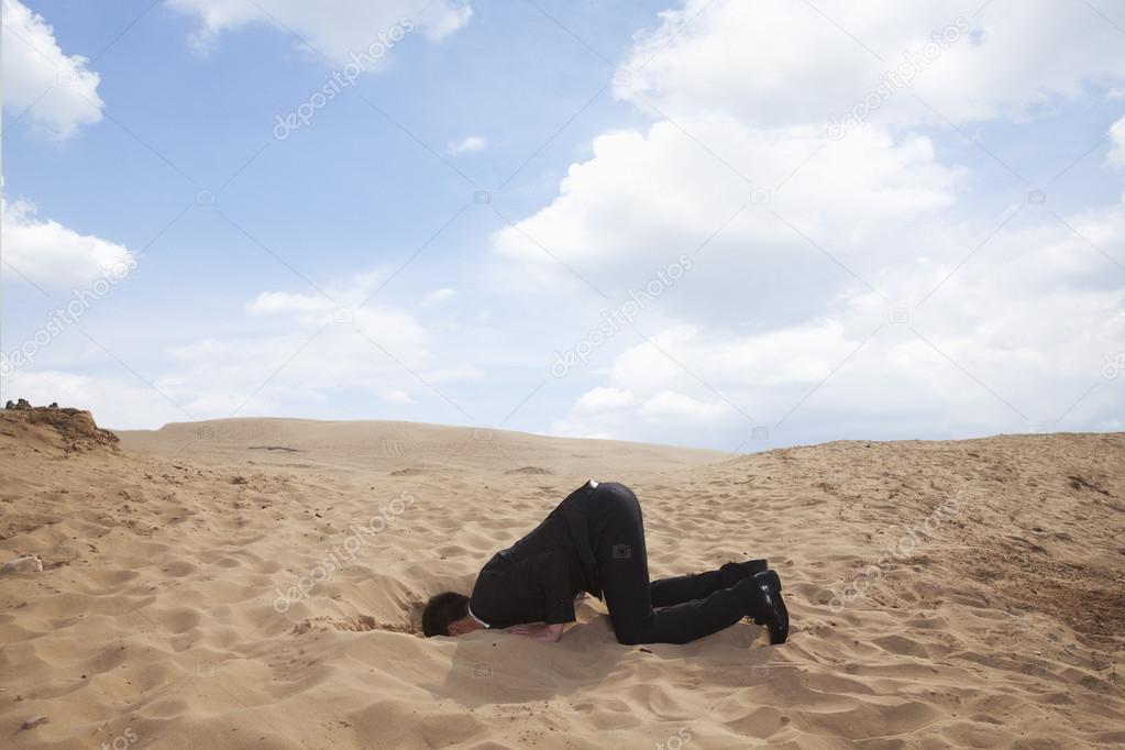 Businessman kneeling with his head in a hole in the sand