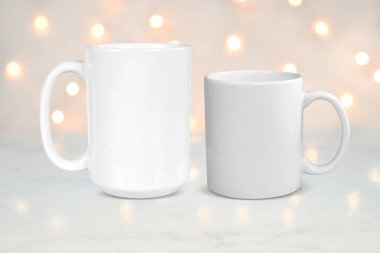 15 oz and 11 oz white coffee mugs resting on a white marble tabletop while white lights shine in the background.