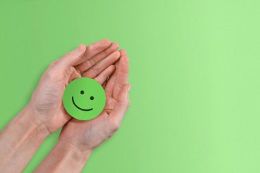 A positive green smiley face being held in a woman's hands against a cheerful green paper background.