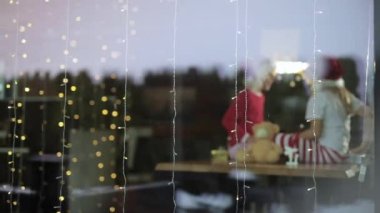 Noel şapkası takan iki çocuk Noel zamanı evde oturuyor ve birbirlerine hediye kutuları veriyorlar.  