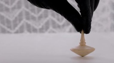 Human hand in black vinyl glove is launching wooden spinning top on a white table.