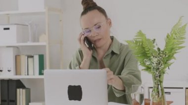 Young female between 30 and 40 years old is speaking on phone in front of pc. 
