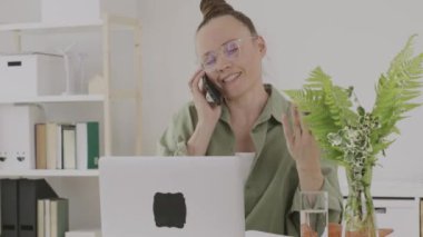 Young female between 30 and 40 years old is speaking on phone in front of pc. 