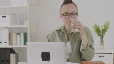 Young female between 30 and 40 years old drinks water in front of pc. 
