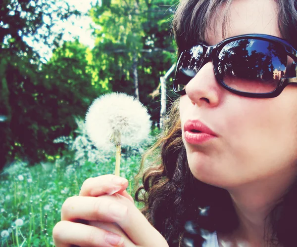 Una mujer guapa con gafas de sol soplando diente de león — Foto de Stock