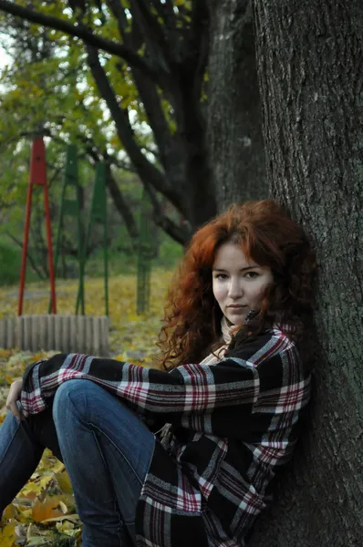 Una hermosa pelirroja con camisa de franela y jeans sentada bajo el árbol de otoño —  Fotos de Stock