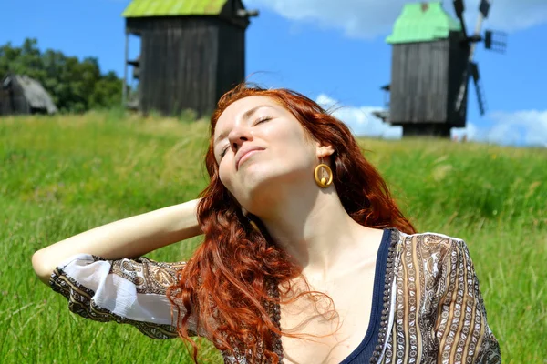 Beautiful young redhead woman smiling and holding her hands in her hair with hear eyes closed on the background of the mills — Stock Photo, Image