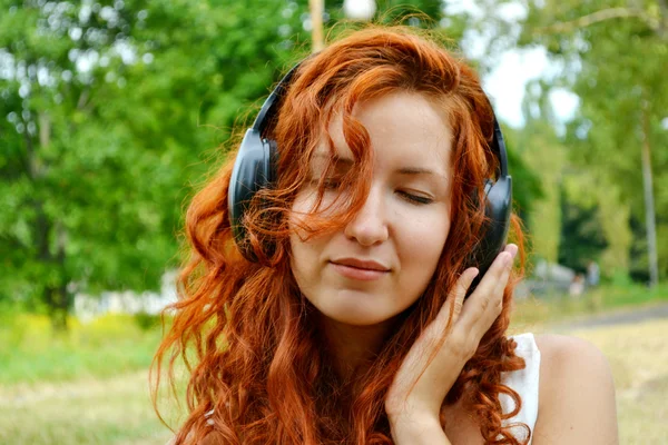 Beautiful redheaded woman in big headphones listening to the music with her eyes closed — Stock Photo, Image