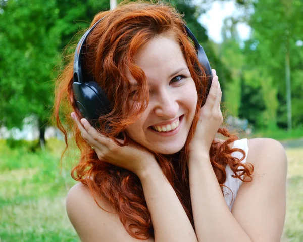 Mujer pelirroja hermosa en grandes auriculares escuchando la música con sonrisa feliz — Foto de Stock