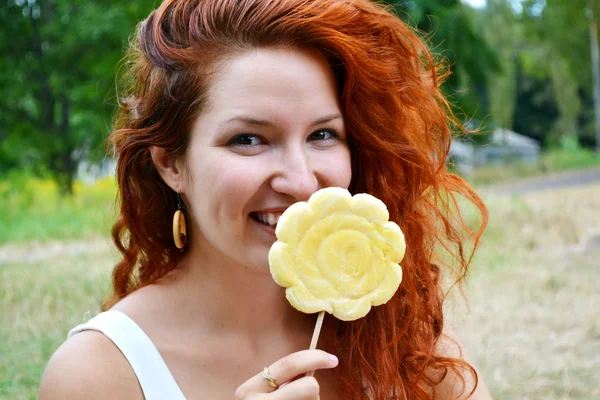 Красива молода руда жінка щасливо посміхається з великим квітковим жовтим льодяником — стокове фото