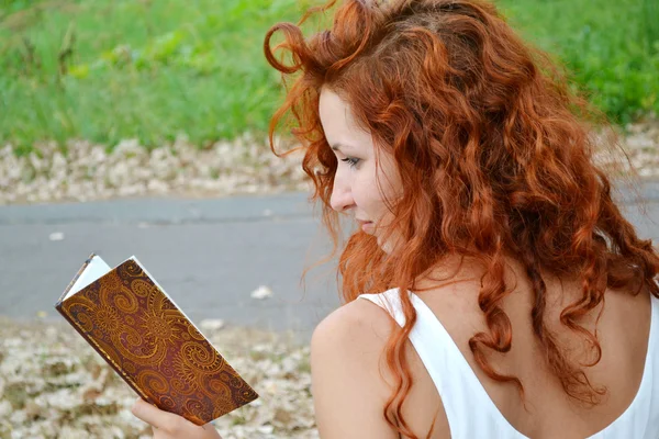 Beautiful redheaded woman with curly hair reading against autumn nature — Stock Photo, Image