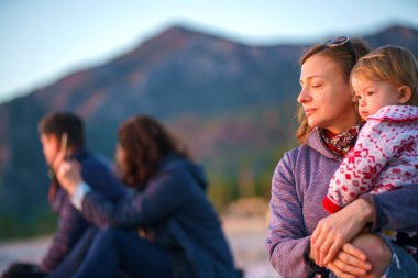 Kucağında bir kızı olan anne, kış denizinin kıyısında bir dağın, ormanın ve iki arkadaşın zemininde oturuyor. Ebeveyn ve çocuk dışarıda, taştan bir sahilde.