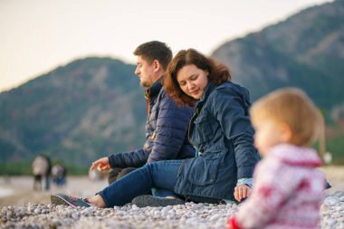 Bir kadın ve bir erkek sahilde kameradan uzakta oturur, bebek kameraya daha yakın oturur - bulanık. Seçici odaklanma. Aile kışın doğada olur. Ailesi ve kızı kış plajında..