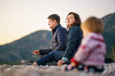 Seçici odaklanma. Ailesi ve kızı kış plajında. Aile kışın doğada olur. Bir kadın ve bir erkek sahilde kameradan uzakta oturur, bebek kameraya daha yakın oturur - bulanık.