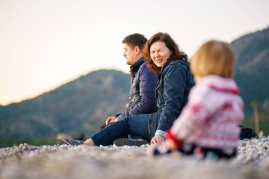 Seçici odaklanma. Aile kışın doğada olur. Ailesi ve kızı kış plajında. Bir kadın ve bir erkek sahilde kameradan uzakta oturur, bebek kameraya daha yakın oturur - bulanık.