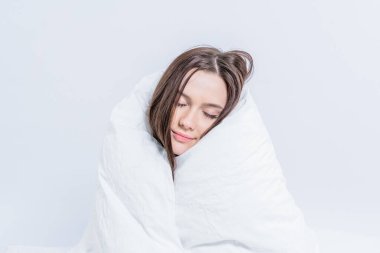 Beyaz bir battaniyeye sarılı güzel bir esmer, yatağa oturur. Sıcak bir battaniyeyle sıcak tutun. Tembellik.