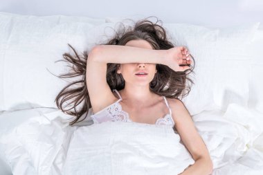 Esmer, beyaz bir yatakta uyuyor. Yatak odası. İyi sabahlar. Rahatlama. Pijamalı tatlı bir kız yatağa uzanmış, gözlerini eliyle kapatıyor..