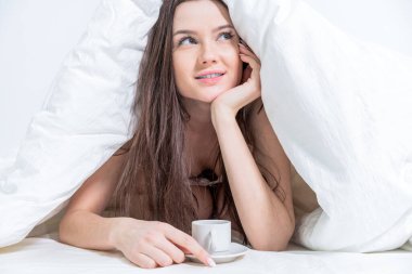 Bir kadın bir fincan kahvenin yanında bir battaniyenin altına uzanır. Lezzetli bir kahvaltı. Esmer, elinde sıcak bir içecekle bir fincan tutuyor. Beyaz yatak odası. Günaydın..
