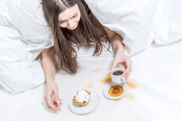 The girl is indignant, spilled coffee on the bed. The brunette lies in bed, next to her is a cupcake, a baguette in her hand and a cup. Breakfast.