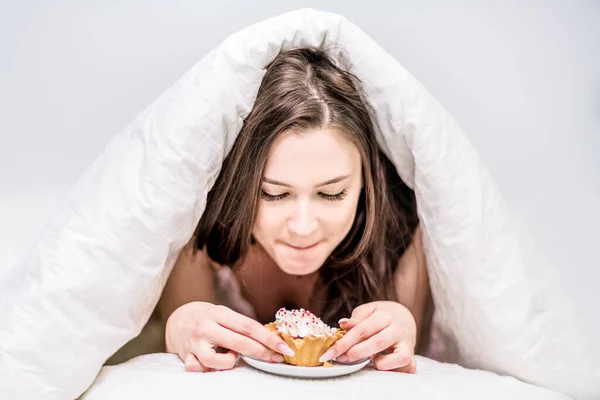 可愛い女は蓋の下のベッドの上に寝そべっているカップケーキを食欲をそそる ベッドで朝食 デザートだ カロリー — ストック写真
