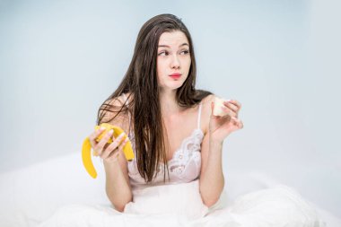 Esmer, meyveyle kahvaltı yapıyor. Kız battaniyeye sarılı olarak yatakta oturuyor ve muz yemekten zevk alıyor..