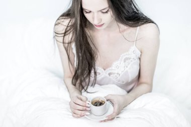 Üzgün kadın yatakta oturup içiyor ve elinde aromatik kahve tutuyor. Lezzetli bir kahvaltı. Esmer, elinde sıcak bir içecekle bir fincan tutuyor. Beyaz yatak odası. Günaydın..