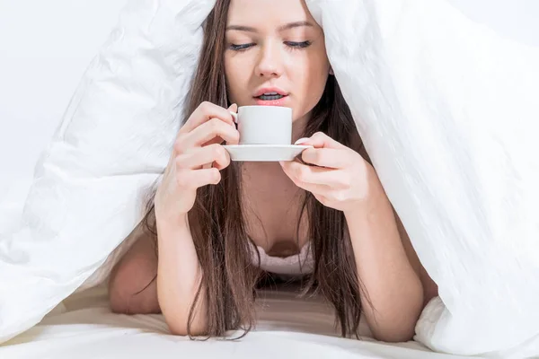 おいしい朝食 ブルネットは熱い飲み物を持っている 女性がカバーの下に横たわっていて コーヒーを見ている おはようございます ホワイトベッドルーム — ストック写真