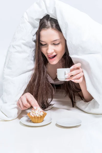 ホワイトベッドルーム おはようございます 女性はベッドに座り 唇を舐め 熱い飲み物と食欲をそそるカップケーキを持っています — ストック写真