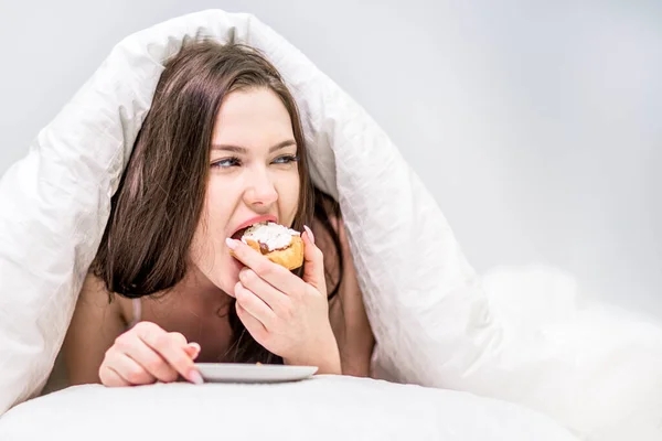 キャリーだ 食欲をそそる可愛い女がカバーの下のベッドの上に寝そべっているカップケーキを食べる デザートだ ベッドで朝食 — ストック写真