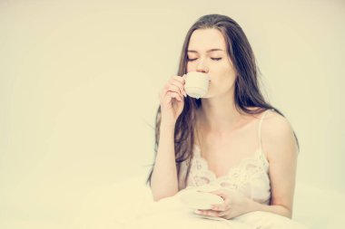 Esmer, elinde sıcak bir içecekle bir fincan tutuyor. Beyaz yatak odası. Tatlı bir kadın yatakta oturuyor ve aromatik kahve içiyor. Lezzetli bir kahvaltı. Günaydın..