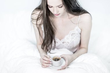 Üzgün kadın yatakta oturup içiyor ve elinde aromatik kahve tutuyor. Lezzetli bir kahvaltı. Esmer, elinde sıcak bir içecekle bir fincan tutuyor. Beyaz yatak odası. Günaydın..
