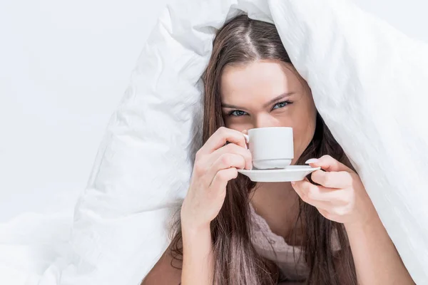 謎の女性がベッドに座って芳香族コーヒーを飲んでいます おいしい朝食 ブルネットは熱い飲み物でカップを持っています ホワイトベッドルーム おはようございます — ストック写真