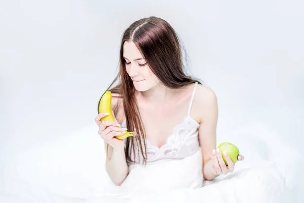 女の子は毛布に包まれ ベッドの上に座ってリンゴを保持し バナナを見て 何を食べるかを選択します ブルネットは果物と朝食を取っています ストックフォト