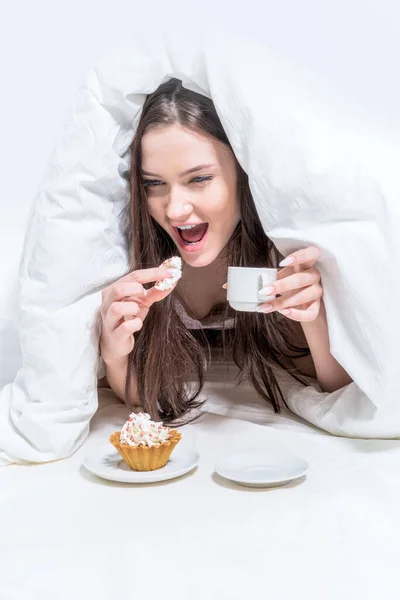 ホワイトベッドルーム おはようございます 面白い女性は 熱い飲み物のカップを保持し 食欲をそそるカップケーキをかむ ベッドに座っています — ストック写真