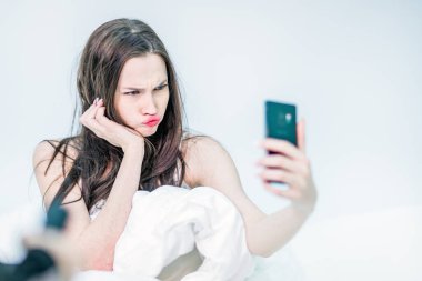 Odasında, beyaz çarşafların arasında elinde akıllı bir telefonla yatan esmer bir kadın. Kız telefon kamerasına surat asıyor, selfie çekiyor..