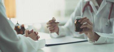 Doktor ve hasta masada otururken bir şey hakkında konuştular. Bir doktor elinde bir şişe ilaç tutarak ne tür bir ilaç olduğunu detaylı bir şekilde açıklıyor..