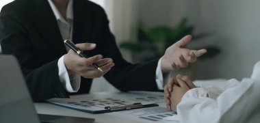 Ekip işkadını işin yapısını anlatıyor ve başka bir çalışan da ihtiyatlı bir şekilde dinliyor. Masadaki belgeler ve ofisteki dizüstü bilgisayarla birlikte.