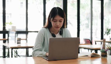 Serious focused Asian student using laptop in kitchen, looking at screen with attention and concentration, watching learning webinar, virtual training, video course, studying from hom