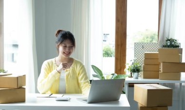 A portrait of Asian woman, e-commerce employee sitting in the office full of packages on the table using a laptop and calculator, for SME business, e-commerce, technology and delivery business