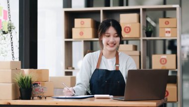 A portrait of a young Asian woman, e-commerce employee sitting in the office full of packages in the background write note of orders and a calculator, for SME business, e-commerce and delivery business