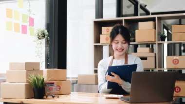 A portrait of a young Asian woman, e-commerce employee sitting in the office full of packages in the background write note of orders and a calculator, for SME business, e-commerce and delivery business