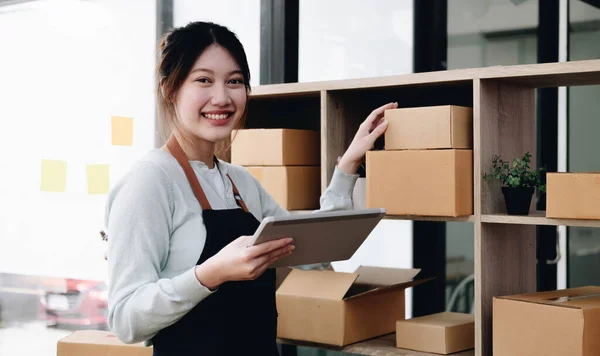Young beautiful happy asian business woman owner of SME online using laptop receive order from customer with parcel box packaging at her startup home office, online business seller and deliver