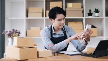Asyalı bir adamın portresi, e-ticaret çalışanı masada hesap makinesi, KOBİ işi, e-ticaret, teknoloji ve dağıtım işi için paket dolu bir ofiste oturuyor.