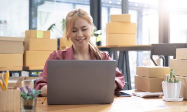 Young beautiful happy asian business woman owner of SME online using laptop receive order from customer with parcel box packaging at her startup home office, online business seller and deliver