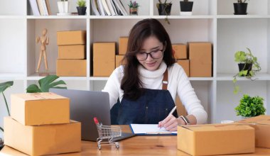 Young beautiful happy asian business woman owner of SME online using laptop receive order from customer with parcel box packaging at her startup home office, online business seller and deliver